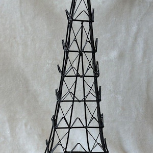 May include: Black metal Eiffel Tower-shaped wine bottle holder. The decorative piece features a detailed lattice design and a pointed top. The holder is approximately 12 inches tall and is designed to hold wine bottles.
