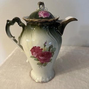 May include: Antique porcelain coffee pot with a dark green lid and handle, and a white body. The pot is decorated with a large pink rose and gold trim. The spout is white with a gold trim. The lid has a small pink rose.