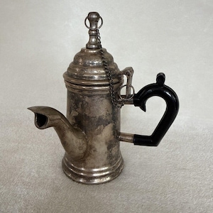 May include: A silver-coloured metal coffee pot with a tarnished finish, featuring a short spout, a domed lid secured by a thin metal chain, and a curved black handle. The cylindrical body has a stepped base and a matching stepped lid design.