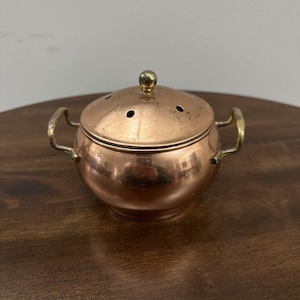 May include: A round copper pot with two brass side handles and a matching domed lid. The lid features a small central brass knob and several small circular ventilation holes. The copper surface has a polished finish with visible metallic tones.