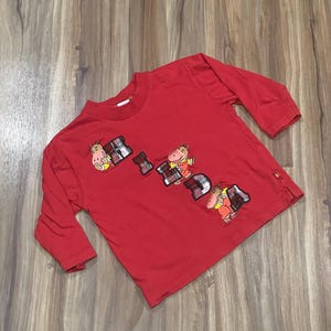 May include: Red long-sleeve shirt with a graphic of the word "Frida" made up of cartoon hippos wearing plaid shirts.