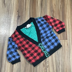May include: A multi-colored plaid cardigan with a V-neck and button closure. The cardigan is made of a soft, comfortable fabric and features a patchwork design with red, blue, and green plaid.