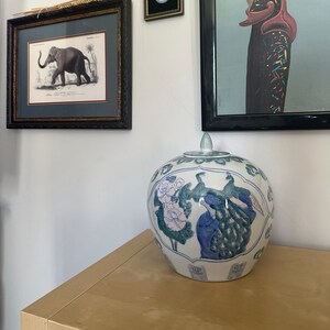 Könnte beinhalten: Eine weiße Keramikvase mit Deckel, verziert mit einem blauen und grünen Design aus Pfauen und Blumen. Die Vase steht auf einer hellbraunen Holzoberfläche.