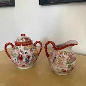 Puede incluir: Un juego de azucarero y jarra de crema de porcelana roja y blanca con un diseño de inspiración japonesa que presenta mujeres con ropa tradicional. El juego tiene un patrón floral y detalles dorados.