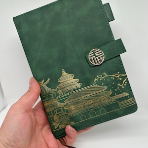 May include: A green leather-bound journal with a gold embossed design of a Chinese temple and a gold double happiness symbol. The journal has a ribbon bookmark and a closure strap.