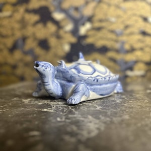 Puede incluir: Una figurita de tortuga de cerámica azul y blanca con un diseño detallado. La tortuga está en posición de caminar con la cabeza levantada y la boca abierta.