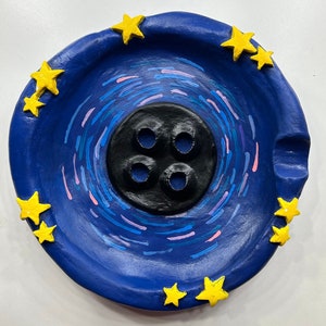 May include: A blue ceramic dish with a black button-shaped center and yellow stars around the edge. The dish has a swirling pattern of blue and pink lines.
