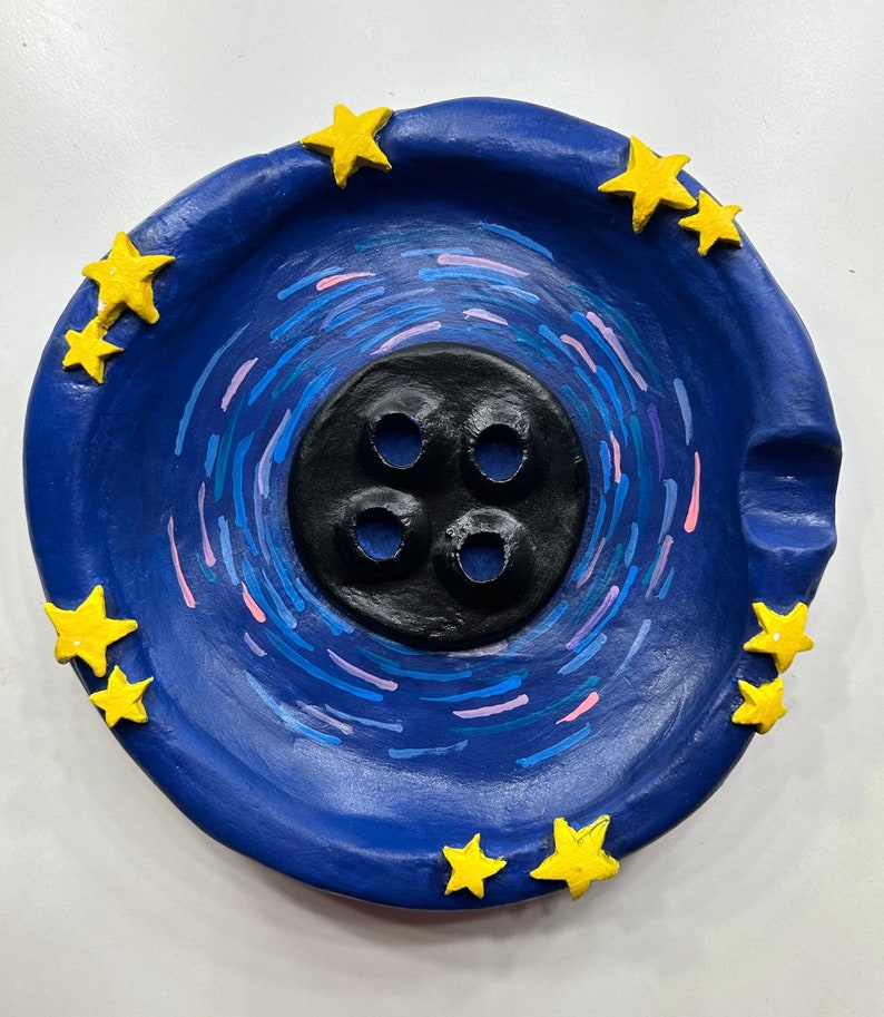 May include: A blue ceramic dish with a black button-shaped center and yellow stars around the edge. The dish has a swirling pattern of blue and pink lines.