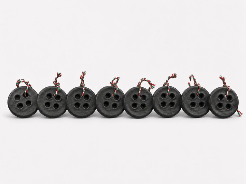 May include: A row of eight circular black buttons, each featuring four central holes. Each button has a small piece of twisted red, white, and green cord tied through the top holes, creating a consistent decorative detail across the set.