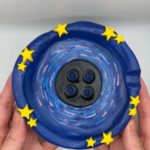 May include: A blue ceramic ashtray with a black center and yellow stars around the rim. The center of the ashtray has a swirling design in blue, pink, and white.