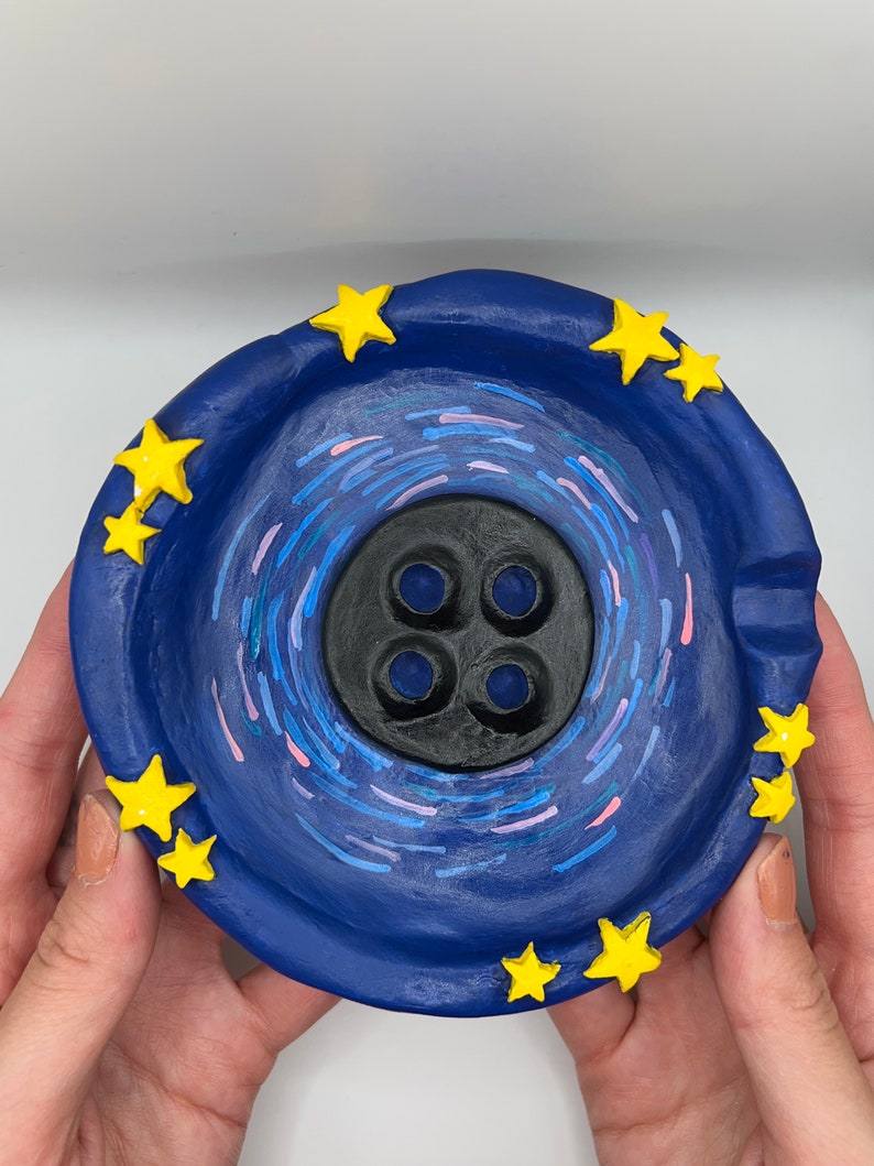 May include: A blue ceramic ashtray with a black center and yellow stars around the rim. The center of the ashtray has a swirling design in blue, pink, and white.