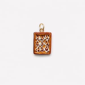 Puede incluir: Un dije colgante rectangular con un dise&ntilde;o de pastel miniatura con corteza dorada y nueve copetes de crema blanca dispuestos en un patr&oacute;n de cuadr&iacute;cula, rematado con una peque&ntilde;a argolla de metal dorado para colgar.