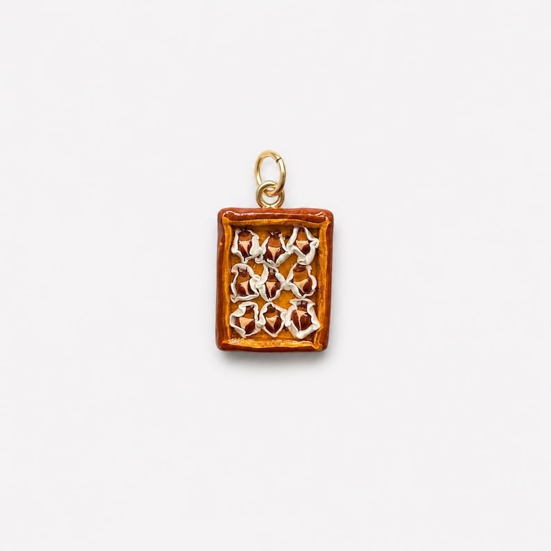 Puede incluir: Un dije colgante rectangular con un dise&ntilde;o de pastel miniatura con corteza dorada y nueve copetes de crema blanca dispuestos en un patr&oacute;n de cuadr&iacute;cula, rematado con una peque&ntilde;a argolla de metal dorado para colgar.