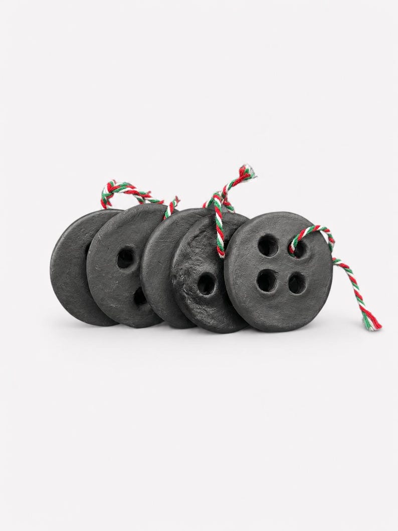 May include: A set of five circular black buttons with a textured, matte finish. Each button features four holes and is threaded with a piece of red, white, and green twisted string. The buttons are arranged in a slightly overlapping horizontal row.