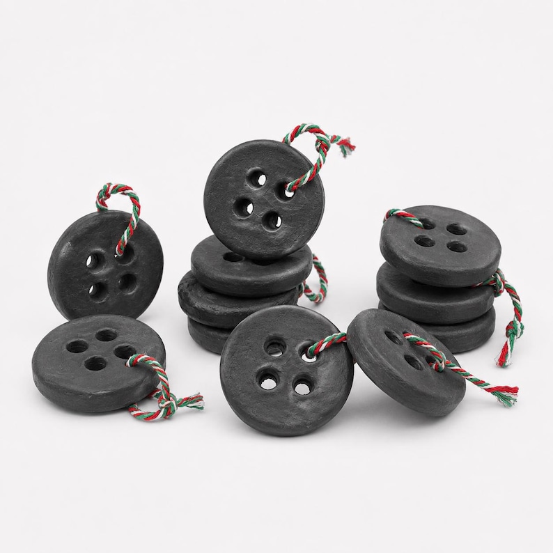 May include: A collection of round, matte black buttons, each featuring four central holes. Each button is threaded with a short piece of twisted red, white, and green string. The buttons are arranged in small stacks and individual pieces.