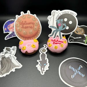 May include: A set of stickers featuring characters and designs from the Netflix show "The Midnight Gospel". The stickers are on a black background and include a pink donut with a cake on top that says "welcome home!" and a blue octopus with a button on its head and a record that says "what's shakin' baby?"