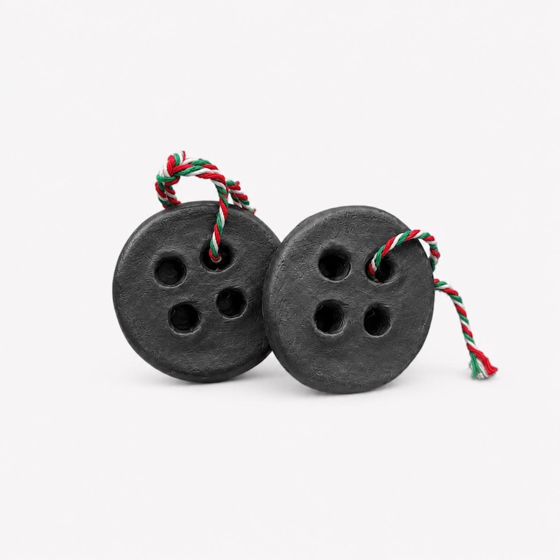 May include: Two round, matte black buttons, each featuring four holes in the center. A piece of twisted red, white, and green cord is threaded through the holes of each button, creating a small loop extending from the top of the circular objects.