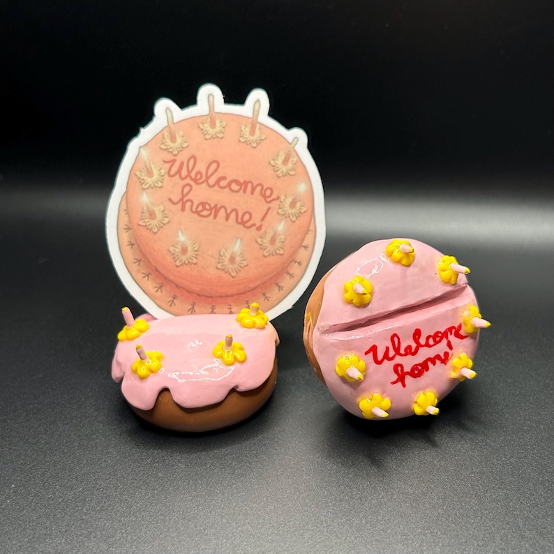May include: Three pink and brown cake-shaped decorations with yellow frosting and candles. The larger decoration has the words "welcome home!" written on it. The smaller decorations have the words "welcome home" written on them.