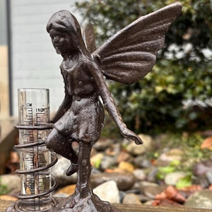 May include: A cast iron fairy rain gauge with a clear glass tube and a metal spiral. The gauge is decorated with a fairy with wings and a dress. The fairy is standing on a base with a textured surface. The gauge is sitting on a wooden surface.