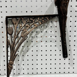 May include: A dark bronze-colored metal shelf bracket featuring an ornate, cast design of tulip flowers and slender stems. The bracket has a right-angle shape with a decorative, curved support arm that connects the vertical and horizontal sections.
