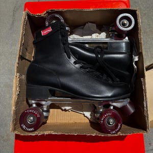 May include: Black roller skates with red wheels and the text "Chicago Urethane" on the wheels. The skates are in a cardboard box on a red surface. The skates have black laces and a high-top design.