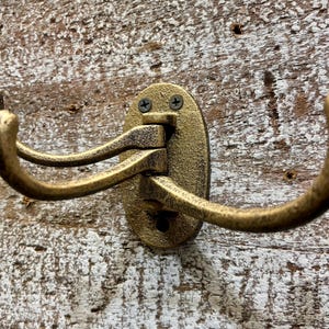 May include: A gold-toned metal coat hook with three hooks. The hook is mounted on a wooden wall.