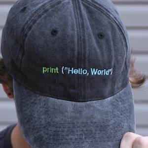Peut inclure: Une casquette de baseball en jean délavé gris anthracite avec le texte brodé print ("Hello, World") sur le panneau avant en fil vert citron et bleu clair. La casquette présente une construction à six panneaux avec une visière courbée.