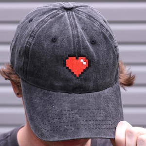 May include: A charcoal grey baseball cap with a distressed, washed-out finish. The front panel features an embroidered pixelated heart design in vibrant red with a small white square accent, mimicking a classic 8-bit video game health icon.