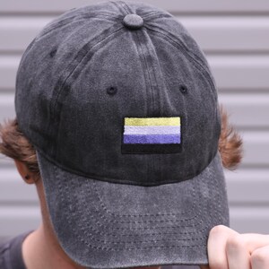 Può includere: Un cappellino da baseball grigio antracite con una finitura slavata e vissuta. Al centro del pannello frontale si trova una toppa rettangolare ricamata che raffigura la bandiera del pride non binario, con strisce orizzontali gialle, bianche, viola e nere.