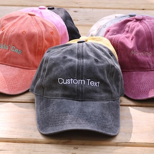 May include: A collection of six baseball caps in various colors including coral, pink, black, yellow, gray, and maroon. Each cap features embroidered text on the front, with the central dark gray cap displaying the words Custom Text.