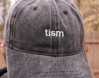 May include: A charcoal gray baseball cap with a washed, distressed finish. The front panel features the word tism embroidered in white lowercase letters. The cap has a curved brim, a six-panel construction, and visible stitching along the seams.