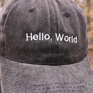 Puede incluir: Una gorra de béisbol gris oscuro con un mensaje bordado en blanco que dice "Hello, World".