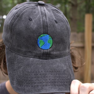 Puede incluir: Una gorra de b&eacute;isbol negra con un globo terr&aacute;queo bordado en azul y verde en la parte delantera.