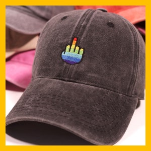 Puede incluir: Una gorra de béisbol de algodón negro, desgastada, con un dedo medio arcoíris bordado en la parte delantera.