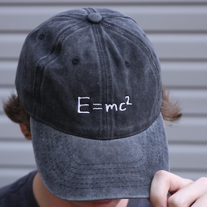 May include: A charcoal grey baseball cap with a distressed, washed-out finish. The front of the cap features the white embroidered text E=mc2 centered above the brim. The cap has a classic six-panel construction with a button at the top center.