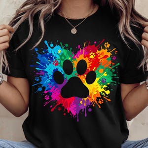 May include: A black short-sleeved t-shirt featuring a large, central graphic of a black animal paw print surrounded by a vibrant, multi-coloured paint splatter design arranged in the shape of a heart, with small paw print accents throughout.