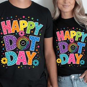 May include: A black t-shirt featuring the text "HAPPY DOT DAY" in large, multi-colored, polka-dotted letters. The design includes a central rainbow-colored dotted circle with a smiley face, surrounded by small colorful dots and flower icons.
