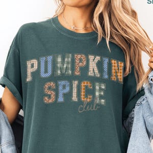 Könnte beinhalten: Ein dunkelblaugrünes Kurzarm-T-Shirt mit dem Schriftzug PUMPKIN SPICE club in Blockschrift. Die Buchstaben haben verschiedene Muster wie Karos, Punkte und Blumen in Blau-, Grün-, Braun- und Orangetönen. Das T-Shirt wird mit einer hellblauen Jeansjacke getragen.