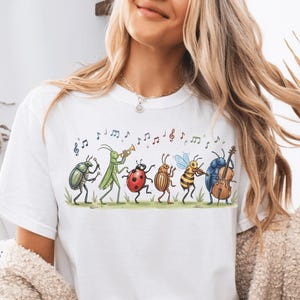 Op de afbeelding: Een wit T-shirt met korte mouwen en een kleurrijke afbeelding van diverse insecten die muziekinstrumenten bespelen, waaronder een kever met trommels, een sprinkhaan met een trompet, een lieveheersbeestje, een bij met een viool en een cello.