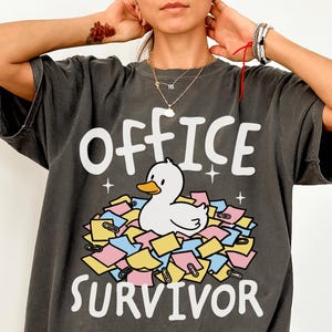 May include: A charcoal grey short-sleeve t-shirt featuring a graphic of a white duck sitting in a pile of colourful sticky notes and paper clips. The text "OFFICE SURVIVOR" is printed in white above and below the duck illustration.