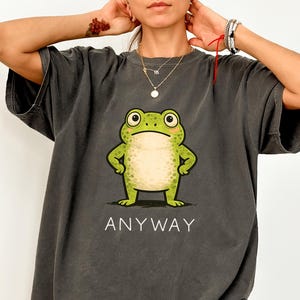 Könnte beinhalten: Pfefferfarbenes T-Shirt mit einem Cartoon-Frosch mit grünem Körper und hellbeige Bauch. Der Frosch hat einen strengen Gesichtsausdruck und verschränkte Arme. Das Wort „ANYWAY“ ist darunter gedruckt.