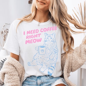 May include: White t-shirt with a blue cat illustration holding a coffee mug with a paw print. The text "I NEED COFFEE RIGHT MEOW" is in pink above the cat. The shirt is a casual, comfortable style.