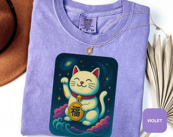 May include: A violet Comfort Colors t-shirt with a Maneki-neko cat graphic. The cat is white with red details, set against a dark blue background with stars and clouds. A gold necklace is draped on the shirt. The text on the coin is in Japanese.