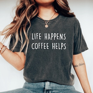 Peut inclure: Un t-shirt gris foncé avec le texte "Life Happens Coffee Helps" imprimé en blanc sur le devant. Le t-shirt est porté par une personne aux cheveux longs et bruns.