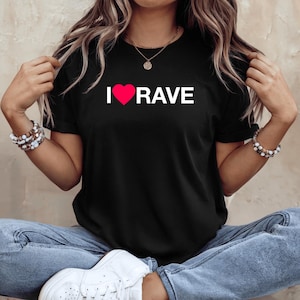 May include: Black t-shirt with the text "I ❤️ RAVE" in white letters and a red heart. The shirt is a crew neck style and is made of a soft material. The shirt is worn by a person wearing blue jeans and white sneakers.