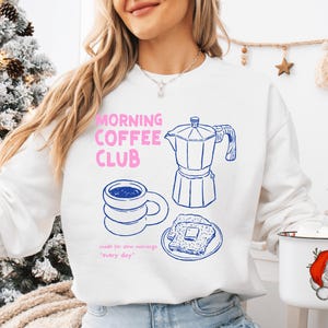 Puede incluir: Sudadera blanca con un gráfico rosa y azul. El diseño incluye el texto "MORNING COFFEE CLUB", una cafetera, una taza de café y tostadas. También se incluye la frase "made for slow mornings 'every day'".
