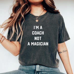 May include: A dark grey cropped t-shirt with the text "I'm a coach not a magician" printed in white. The shirt is being worn by a person with long brown hair.