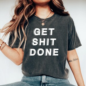 Può includere: T-shirt grigio scuro con la scritta bianca "GET SHIT DONE" in grassetto. La maglietta ha un aspetto vintage e sbiadito. La modella indossa jeans blu e una collana d'oro. Il testo è in grassetto, senza grazie.