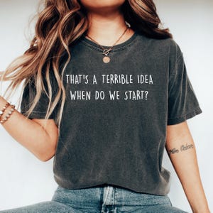 May include: Dark gray t-shirt with the text "THAT'S A TERRIBLE IDEA WHEN DO WE START?" in white. The shirt has a vintage, worn look. The model is wearing a gold necklace and bracelet.
