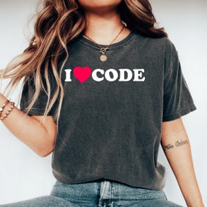 Op de afbeelding: Donkergrijs t-shirt met de tekst "I ❤️ CODE" in witte letters en een rood hart. Het shirt is een casual stijl, waarschijnlijk gemaakt van katoen, en geschikt voor dagelijks gebruik. Het ontwerp is eenvoudig en gedurfd, perfect voor programmeurs en codeurs.